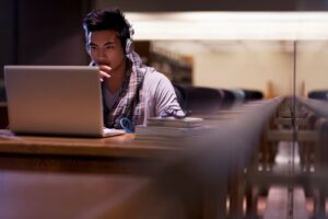 Student wearing headphones and typing on a laptop at a library table with stacked books nearby, focused on his work.