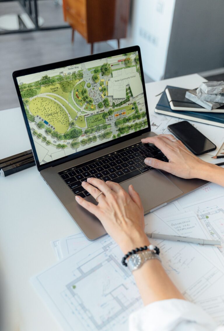 Site plan displayed on a laptop screen, showing green park areas, pathways, and buildings, while someone works nearby.