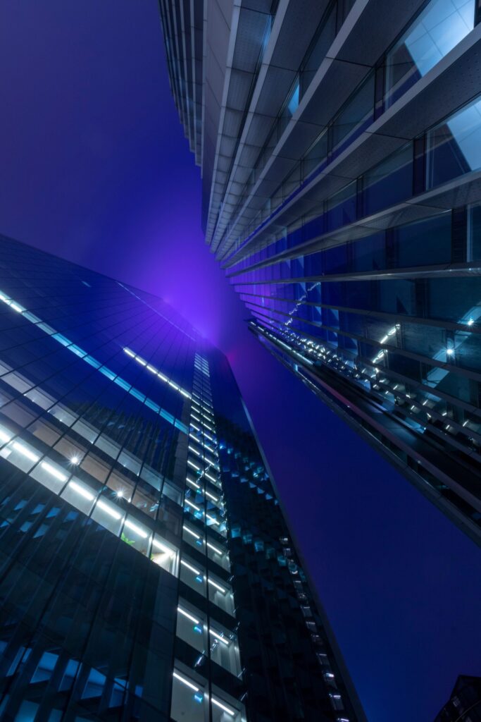 Nighttime view of glass skyscrapers lit in blue and purple, converging toward the center from a low angle.